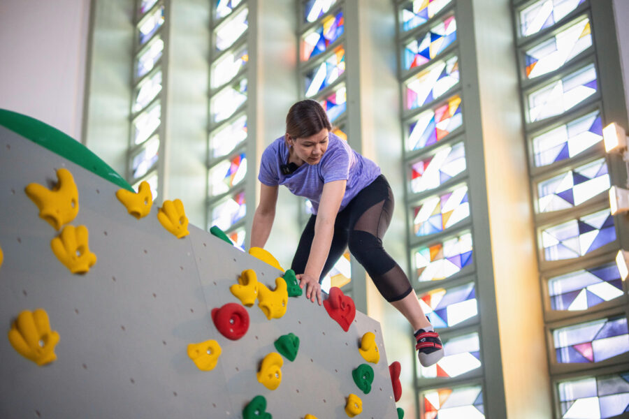Person klettert an einer Wand mit bunten Griffen, bunte Fenster im Hintergrund.