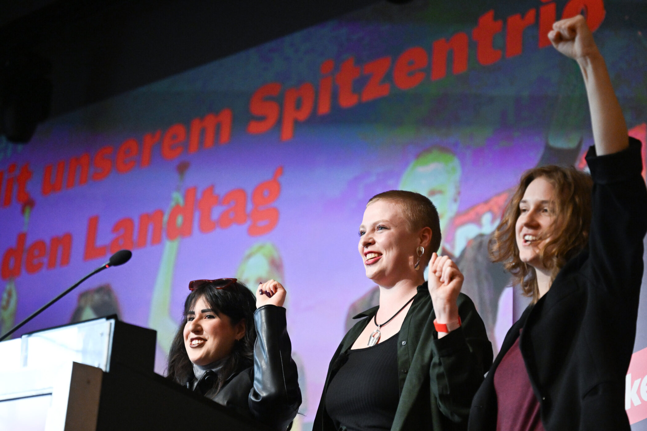Drei Personen auf einer Bühne, heben die Fäuste, Text im Hintergrund.