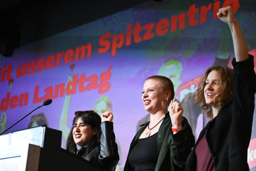 Drei Personen auf einer Bühne, heben die Fäuste, Text im Hintergrund.