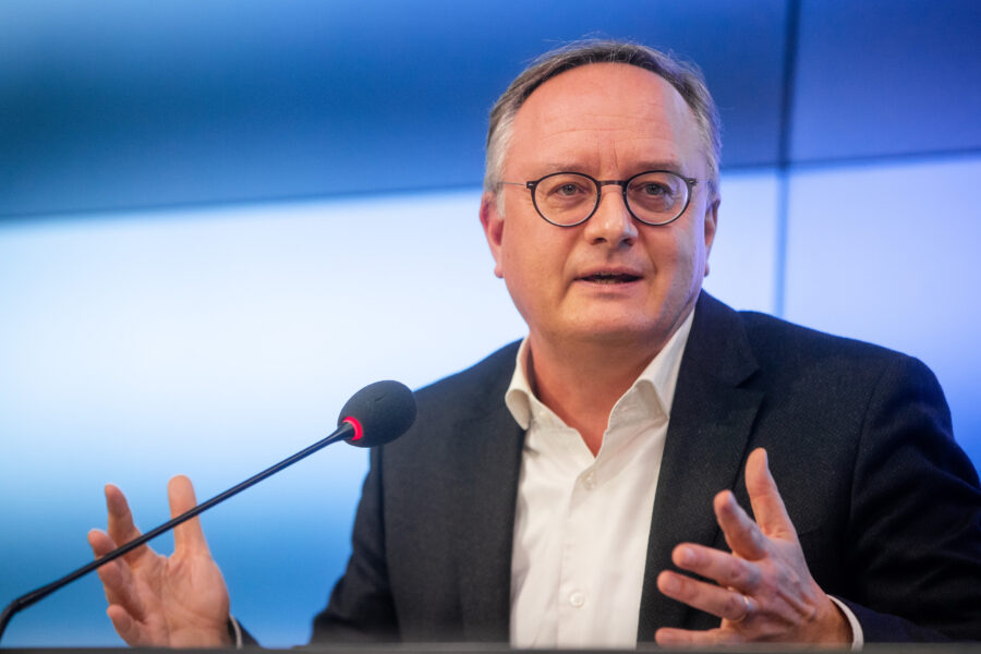 Mann mit Brille spricht in ein Mikrofon, blaue Hintergrundbeleuchtung.