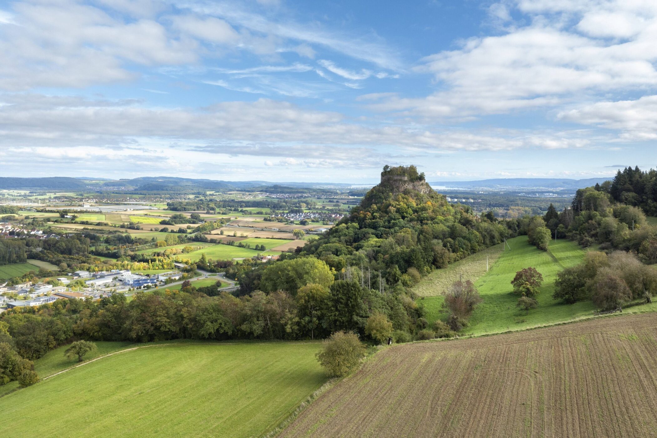 Hügelige Landschaft mit Feldern, Wäldern und einem bewaldeten Hügel unter blauem Himmel.