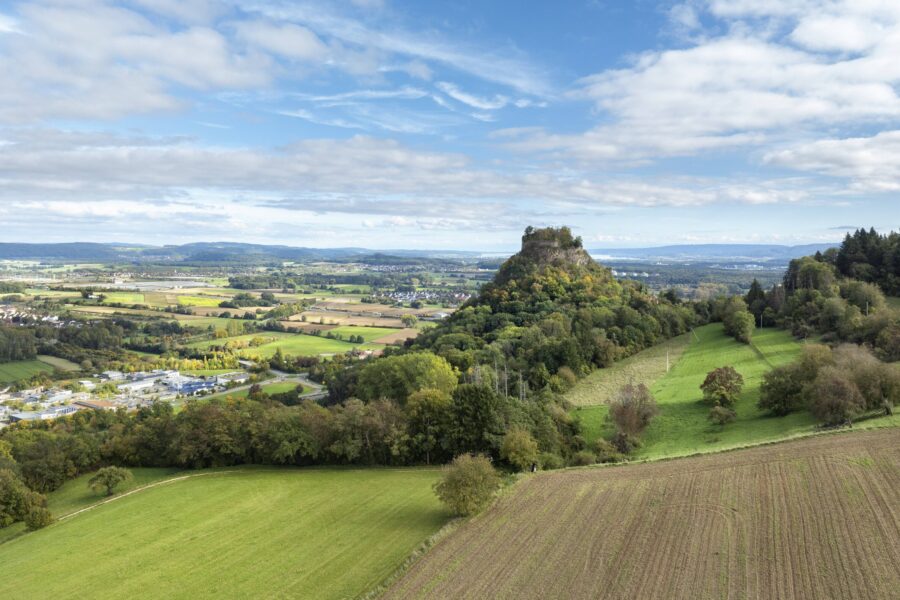Hügelige Landschaft mit Feldern, Wäldern und einem bewaldeten Hügel unter blauem Himmel.