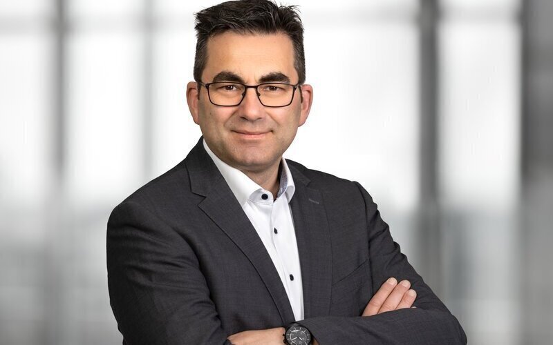 Mann mit Brille im Anzug, Arme verschränkt, vor unscharfem Hintergrund.