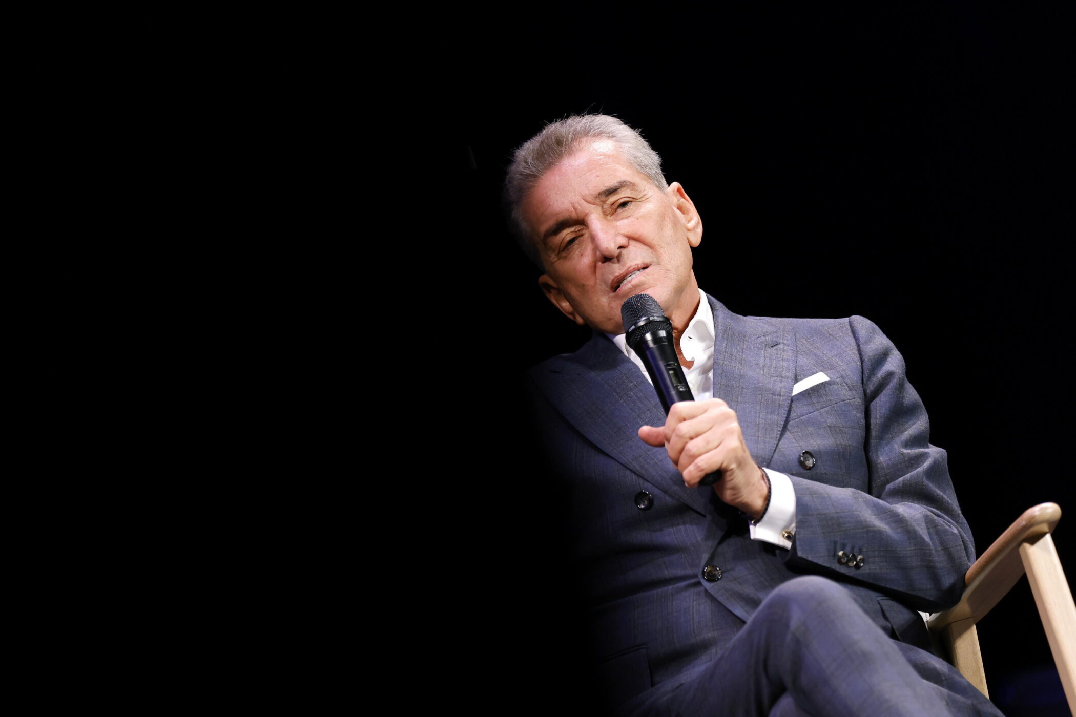 Älterer Mann in Anzug hält Mikrofon, sitzt auf Stuhl vor schwarzem Hintergrund.