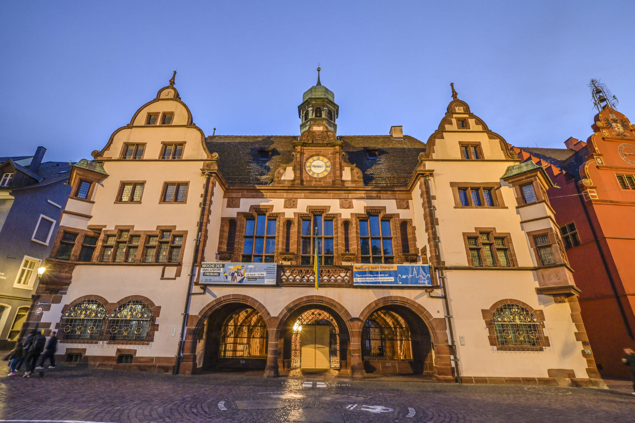 Historisches Gebäude mit Uhrturm, drei Bögen, Bannern und Passanten bei Dämmerung.