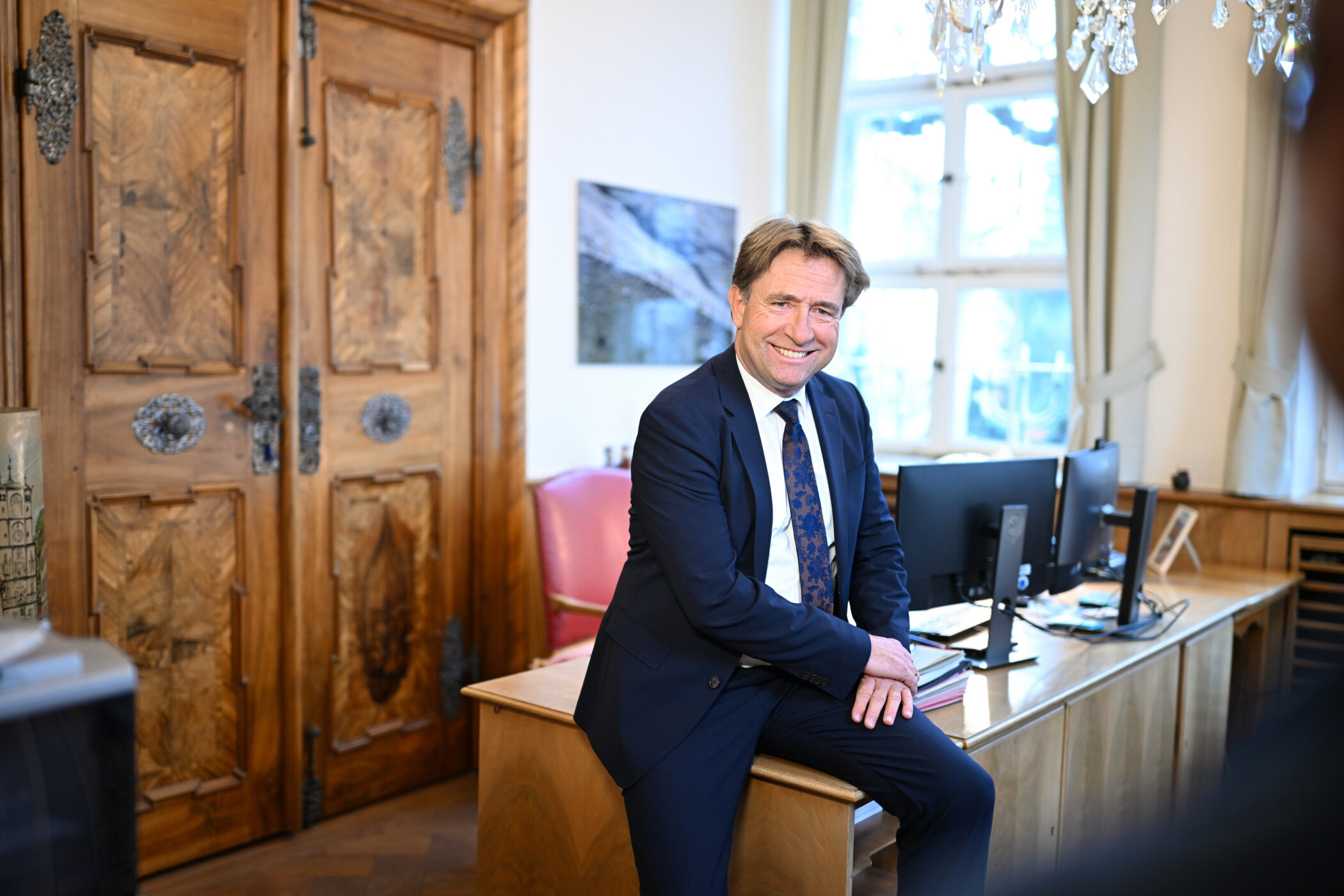 Mann in Anzug sitzt auf Schreibtisch, lächelt, im Büro mit Holztüren und Monitoren.