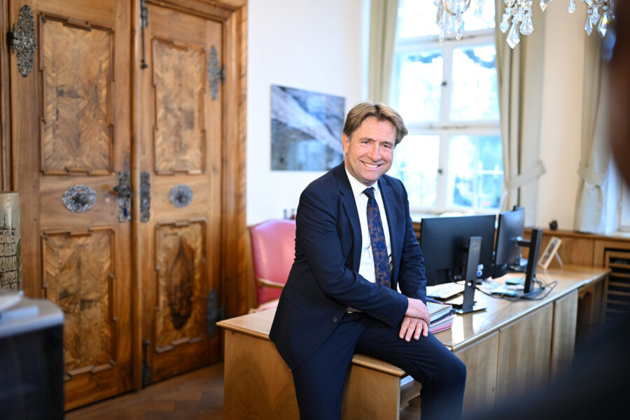 Mann in Anzug sitzt auf Schreibtisch, lächelt, im Büro mit Holztüren und Monitoren.