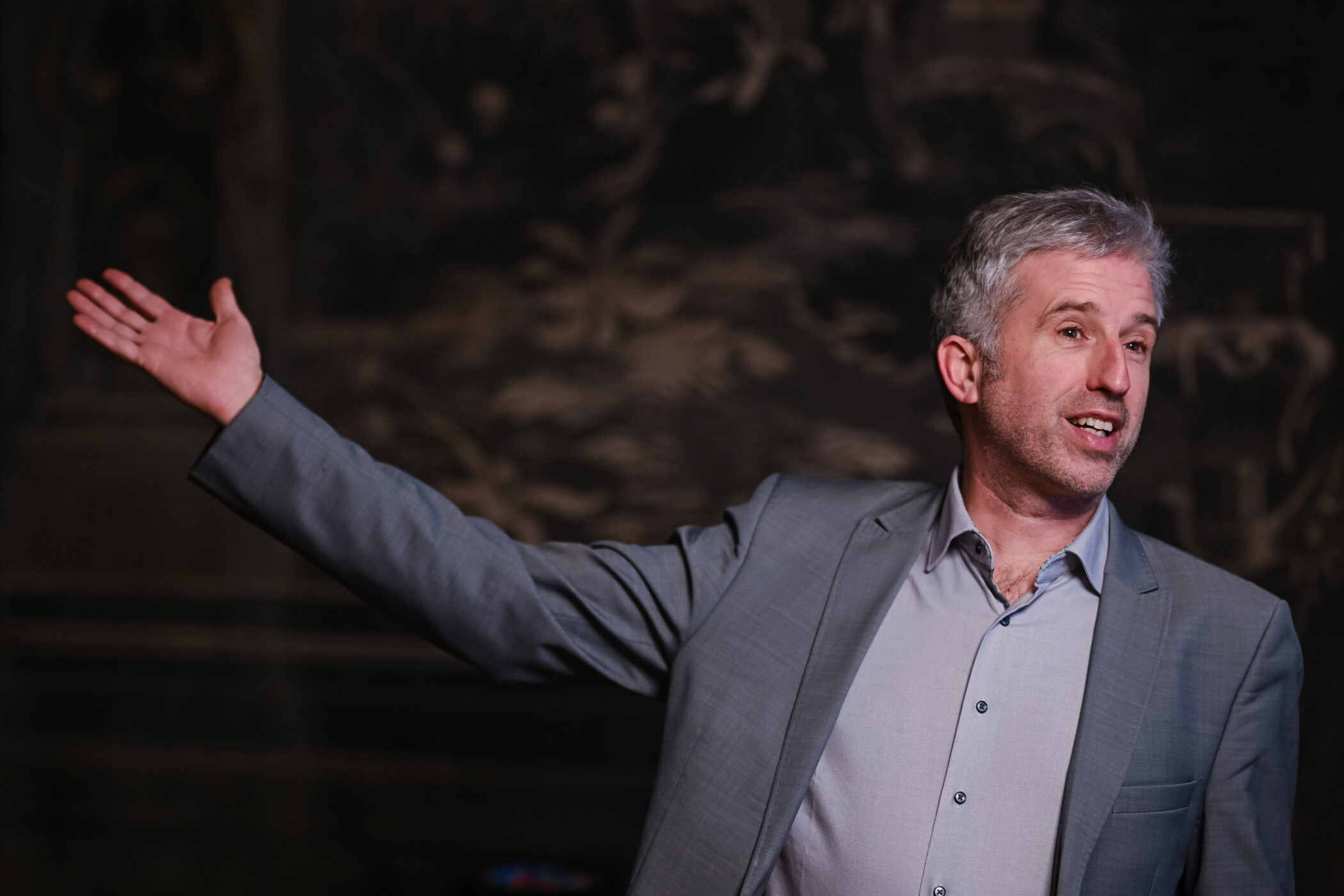 Mann in grauem Anzug gestikuliert mit ausgestrecktem Arm vor dunklem Hintergrund.