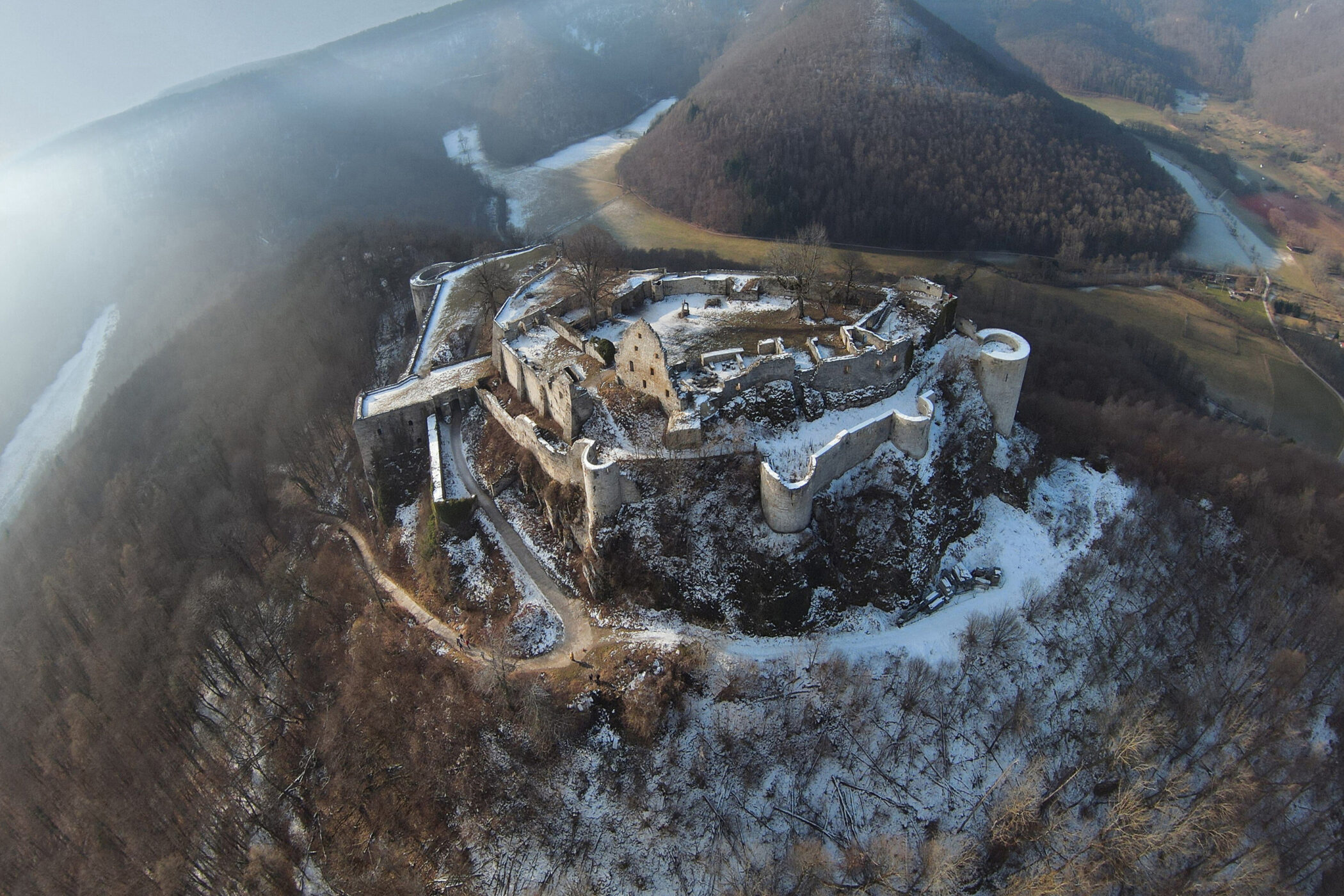 Luftaufnahme einer verschneiten Burgruine auf einem Hügel, umgeben von Wald.