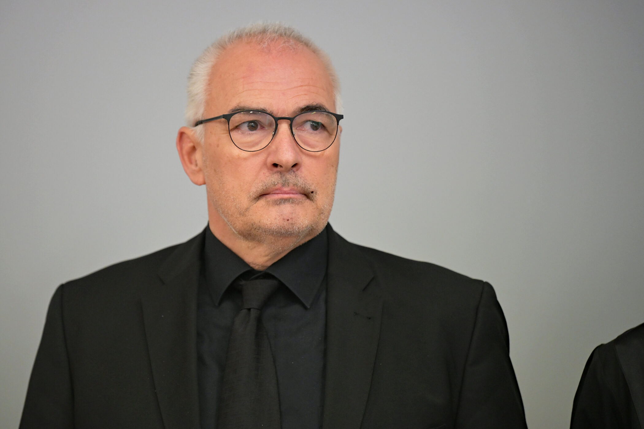 Älterer Mann mit Brille und schwarzem Anzug vor grauem Hintergrund.