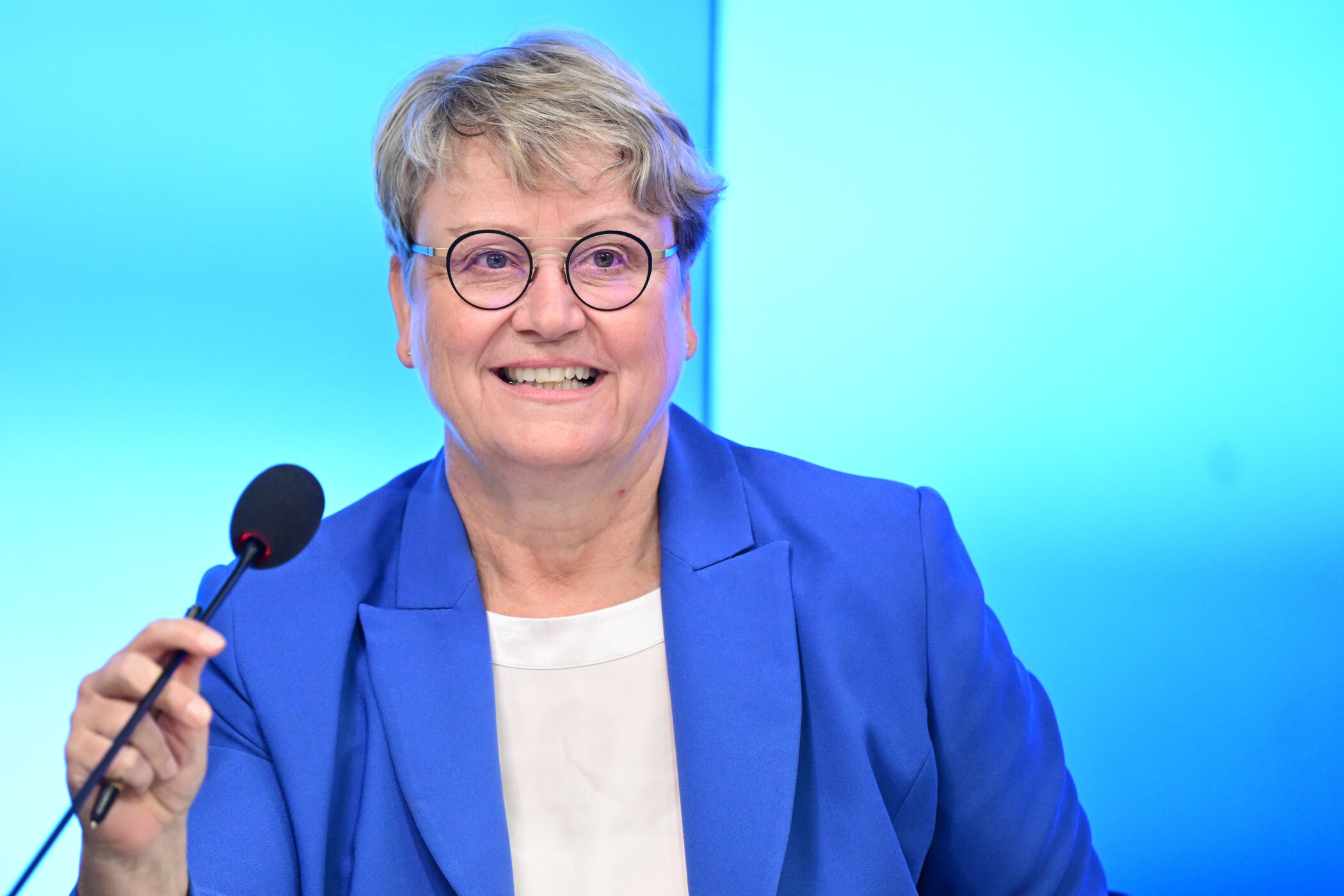 Ältere Person mit Brille und blauem Blazer spricht in ein Mikrofon.