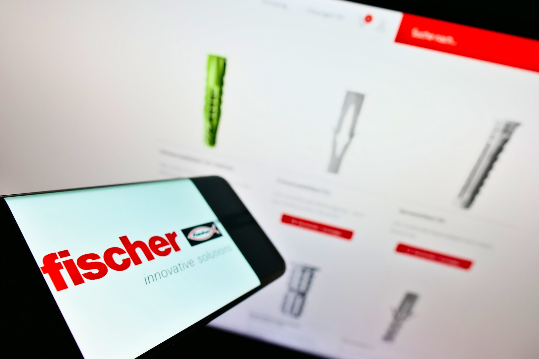 Smartphone mit "fischer" Logo vor unscharfem Bildschirm mit Dübelbildern.