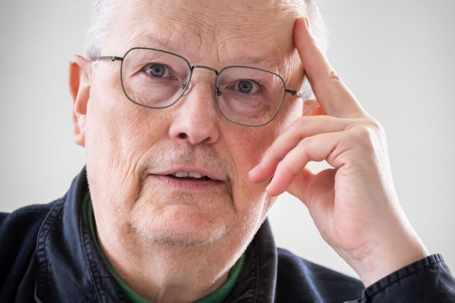 Älterer Mann mit Brille, Hand am Kopf, neutraler Gesichtsausdruck.