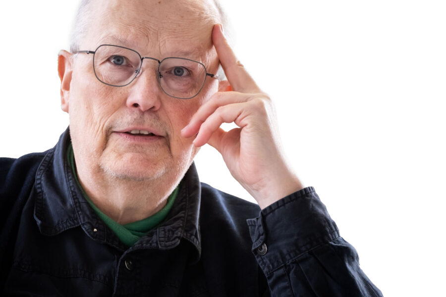 Älterer Mann mit Brille, Hand am Kopf, trägt dunkles Hemd vor weißem Hintergrund.