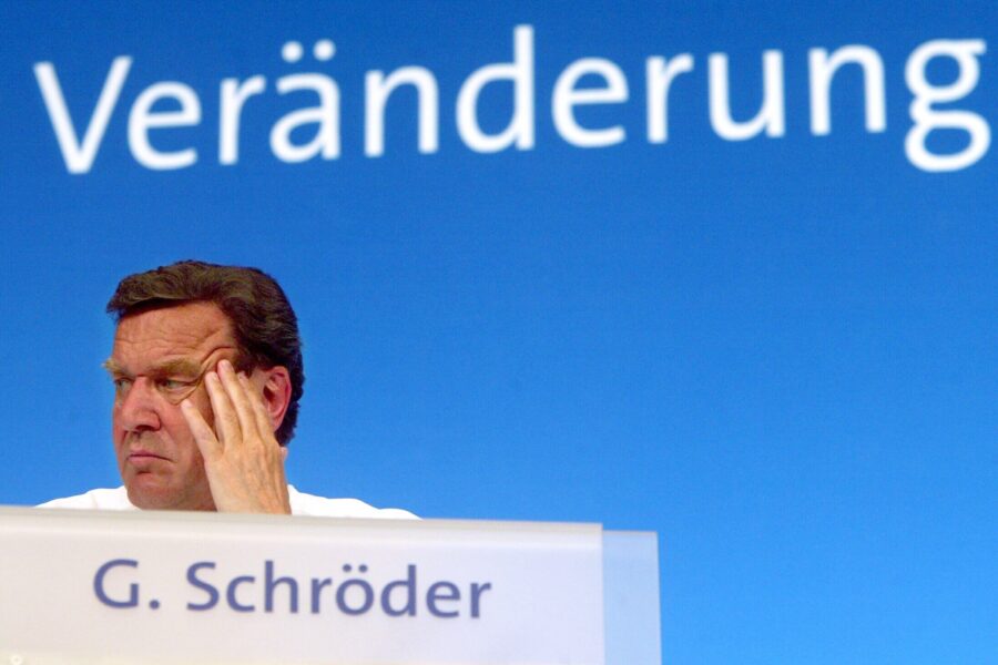 Mann sitzt vor blauem Hintergrund mit dem Wort "Veränderung". Namensschild: G. Schröder.
