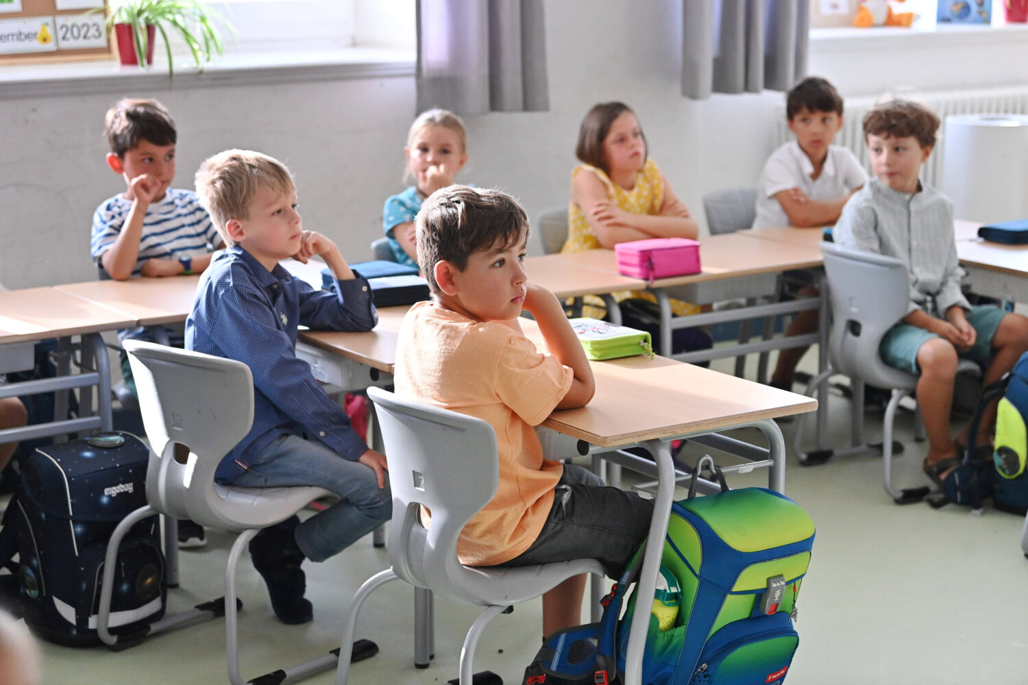 Kinder sitzen an Schultischen und hören aufmerksam zu.