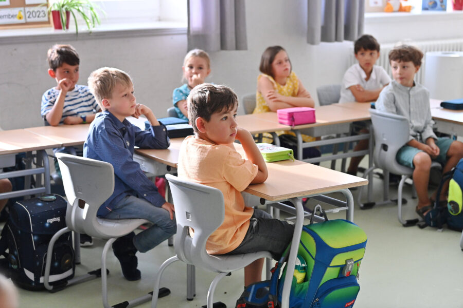 Kinder sitzen an Schultischen und hören aufmerksam zu.