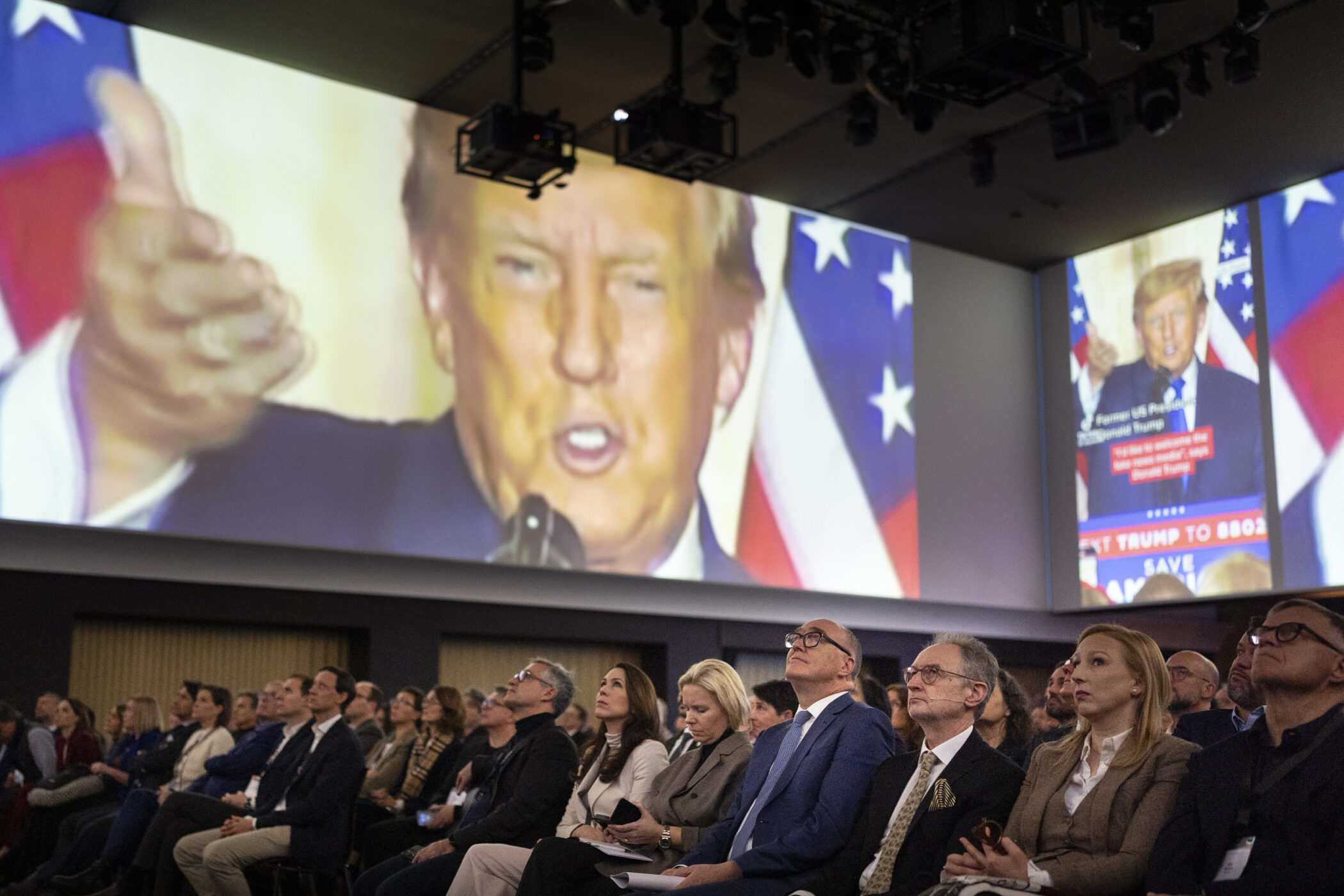 Menschen sitzen in einem Raum, große Projektion eines Mannes im Hintergrund.