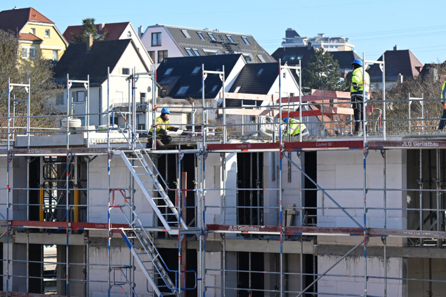 Mehrere Bauarbeiter auf einem Gerüst vor einem Rohbau. Häuser im Hintergrund.