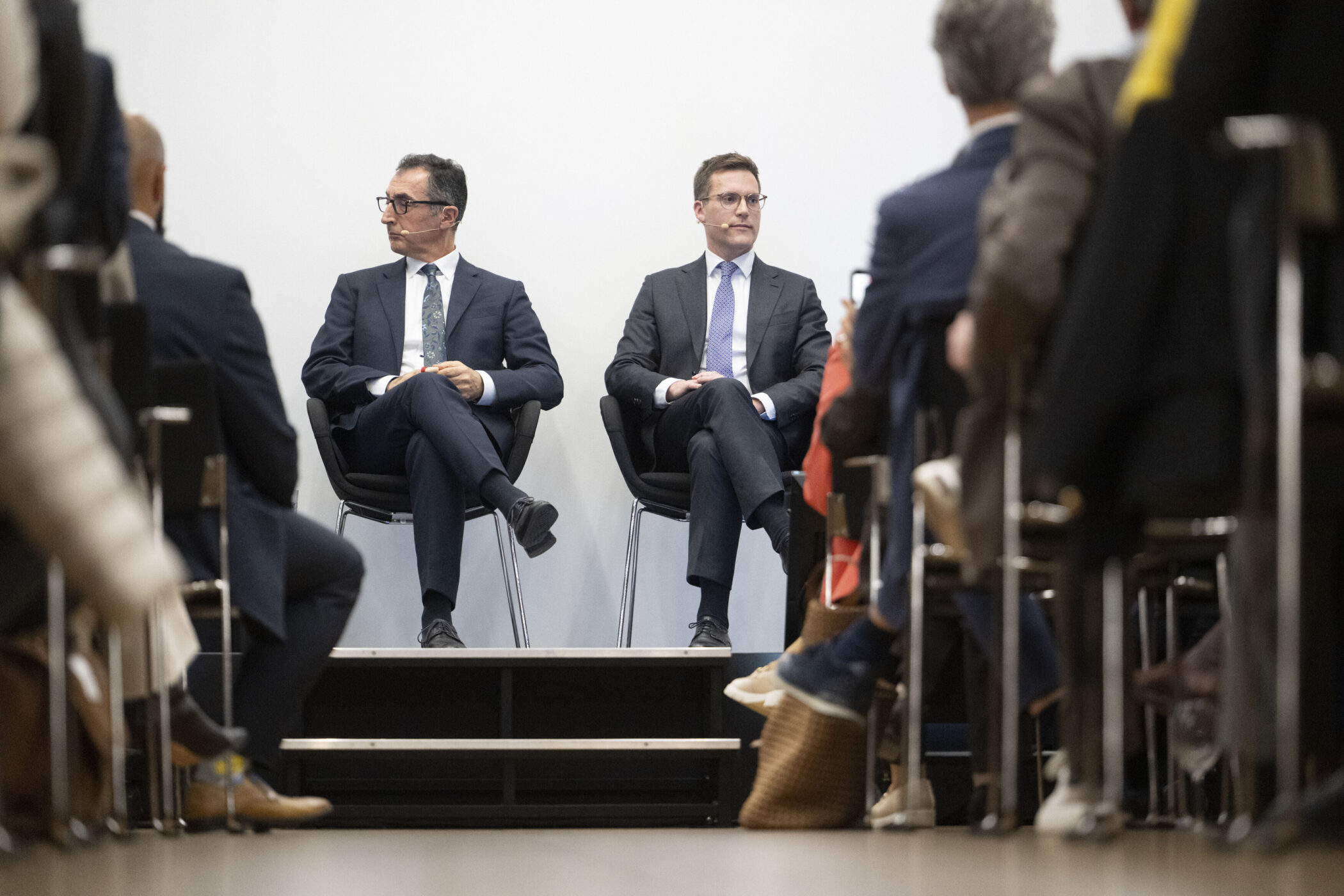 Zwei Männer in Anzügen sitzen auf einer Bühne, Publikum im Vordergrund.