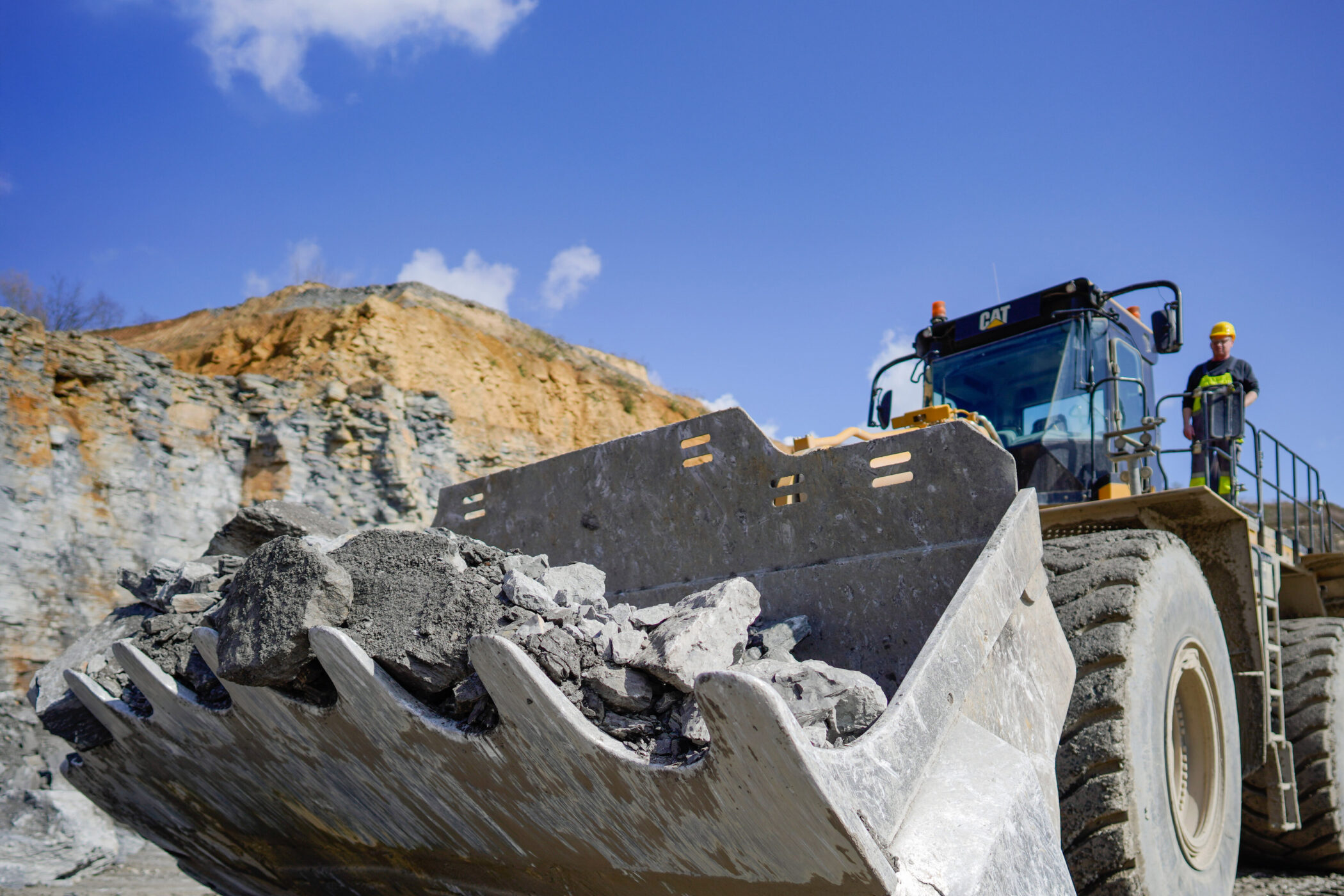 Großer Radlader mit Schaufel voller Steine, zwei Personen mit Helmen daneben.
