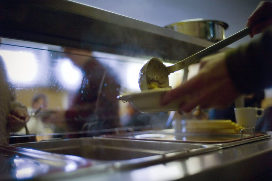 Person schöpft Essen mit Kelle in Schale an Essensausgabe.