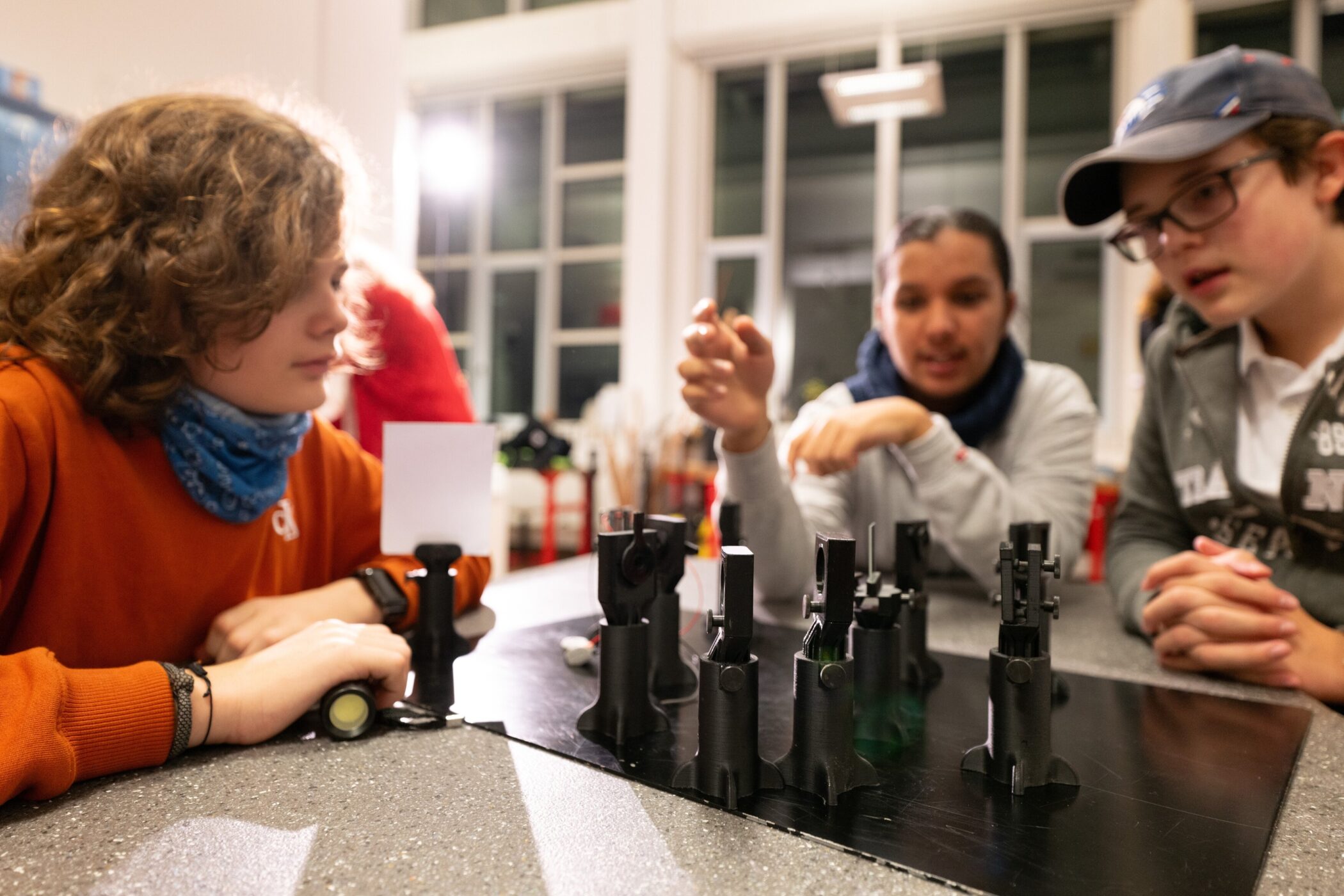 Drei Personen betrachten optische Geräte auf einem Tisch in einem Raum.