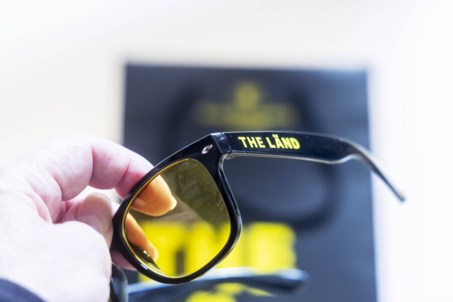Hand hält schwarze Brille mit gelben Gläsern, Aufschrift "THE LAND".