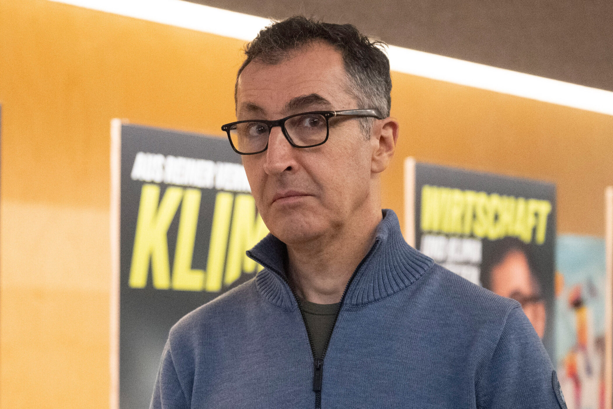 Mann mit Brille und blauem Pullover vor unscharfen Postern im Hintergrund.