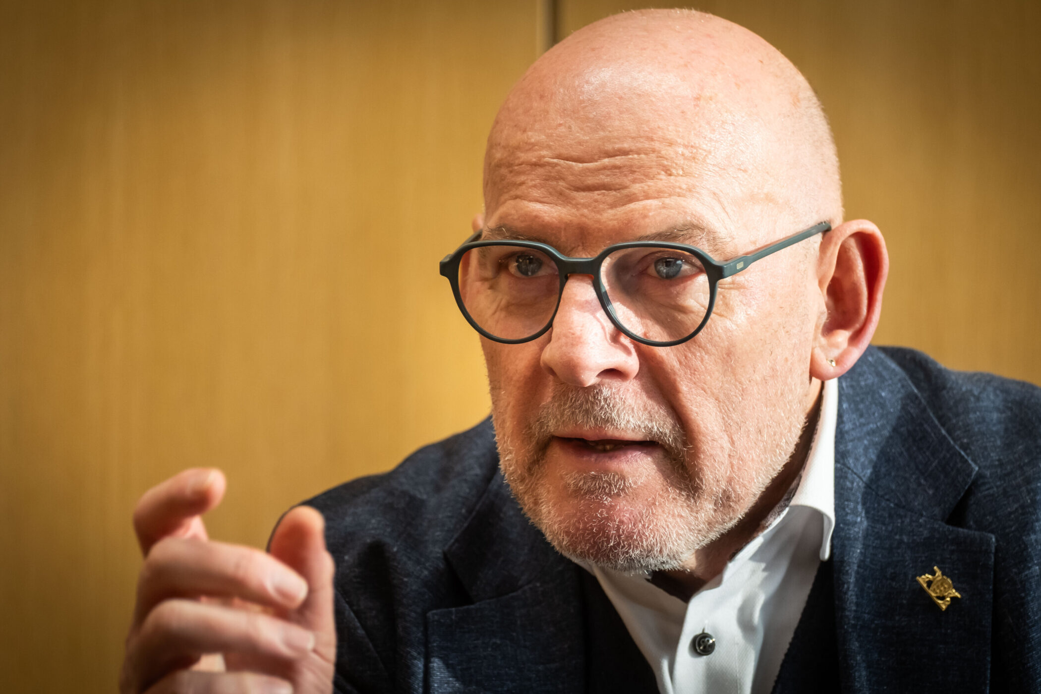 Älterer Mann mit Brille und Anzug spricht, Hand erhoben, Hintergrund unscharf.