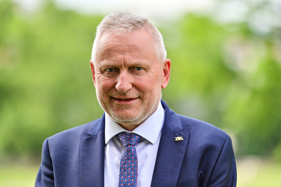 Älterer Mann im Anzug mit Krawatte, lächelnd, unscharfer grüner Hintergrund.