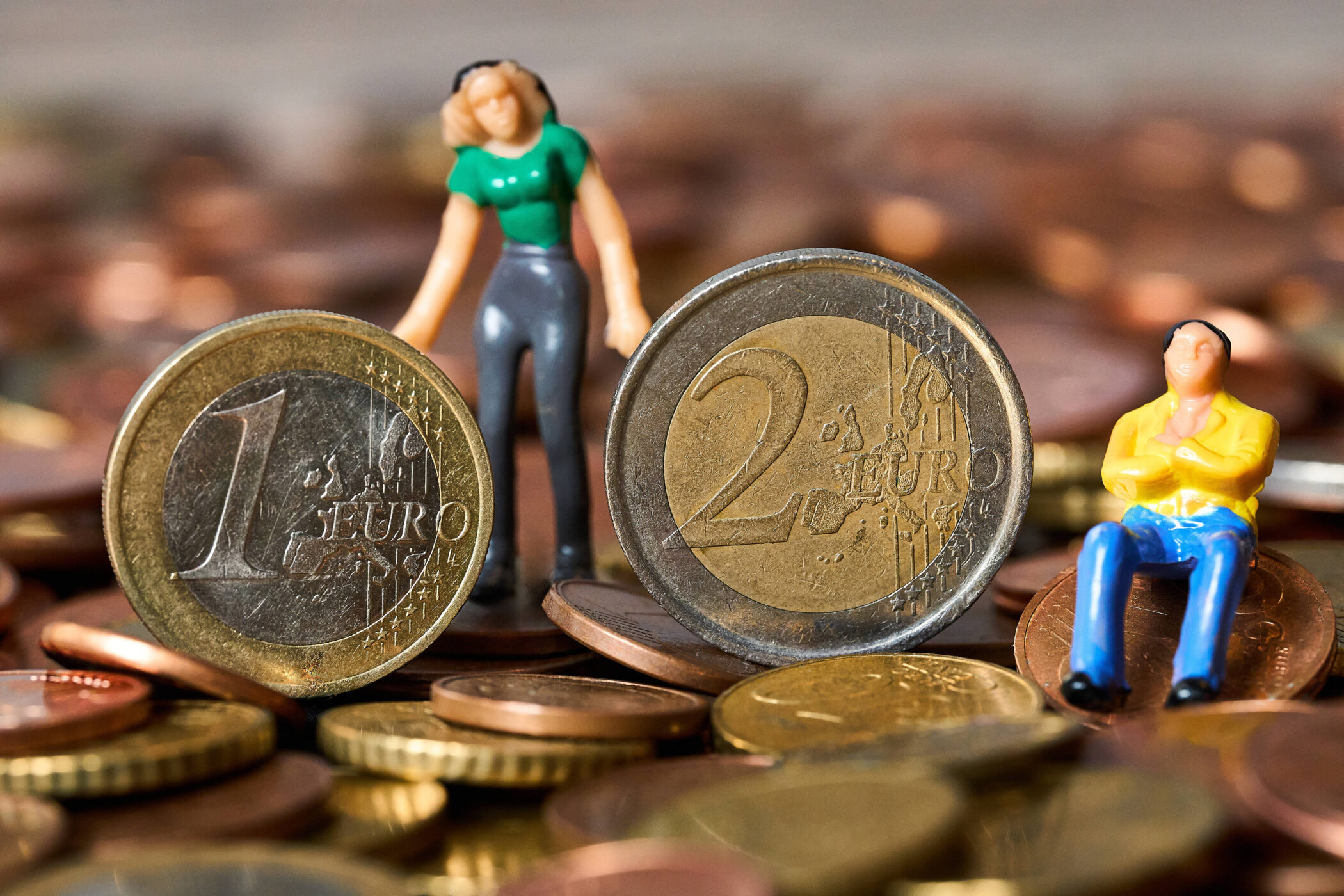 Zwei Miniaturfiguren bei großen 1- und 2-Euro-Münzen auf Haufen Münzen.