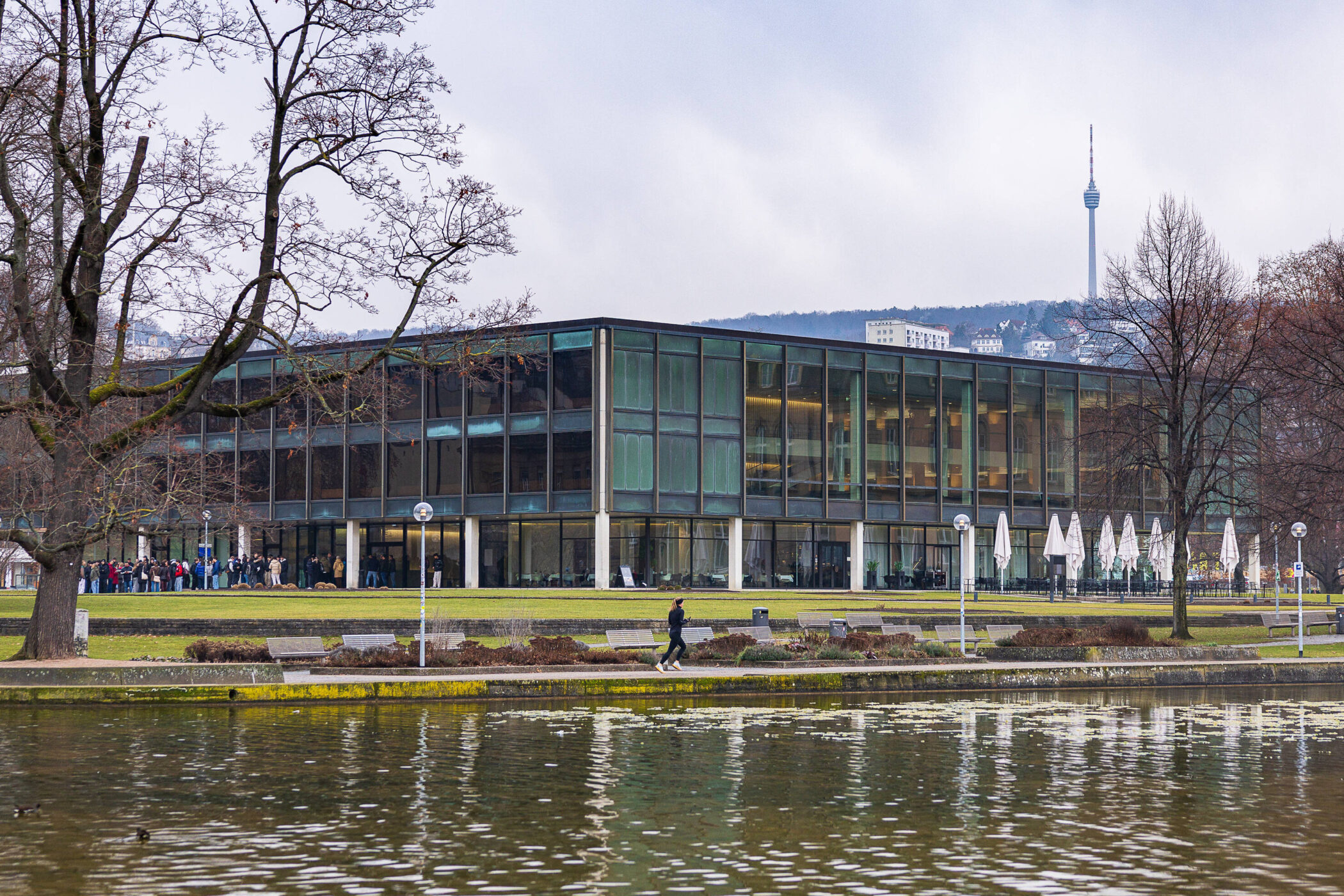 Moderne Glasgebäude am Wasser, Menschen und Bäume im Vordergrund, Turm im Hintergrund.