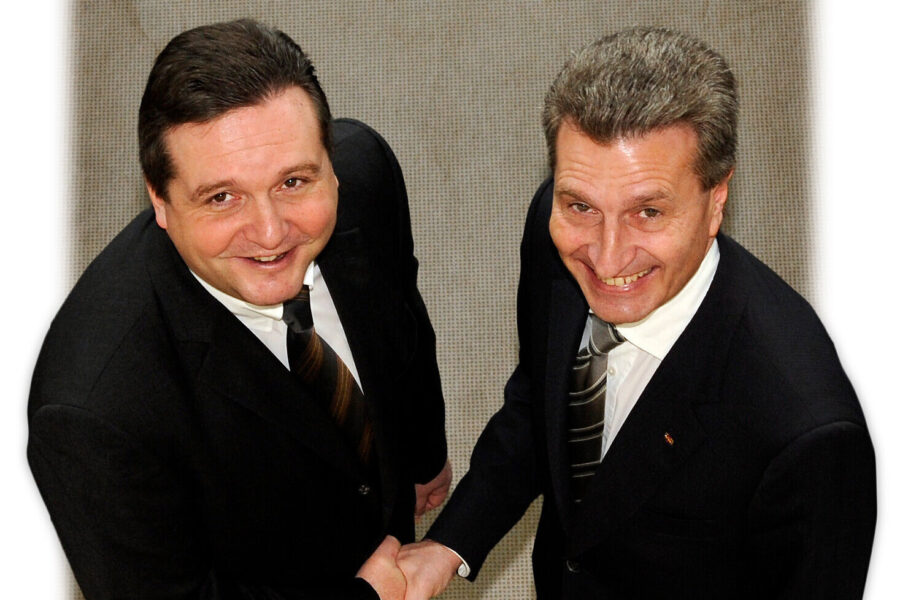 Zwei Männer in Anzügen schütteln sich die Hand und lächeln in die Kamera.