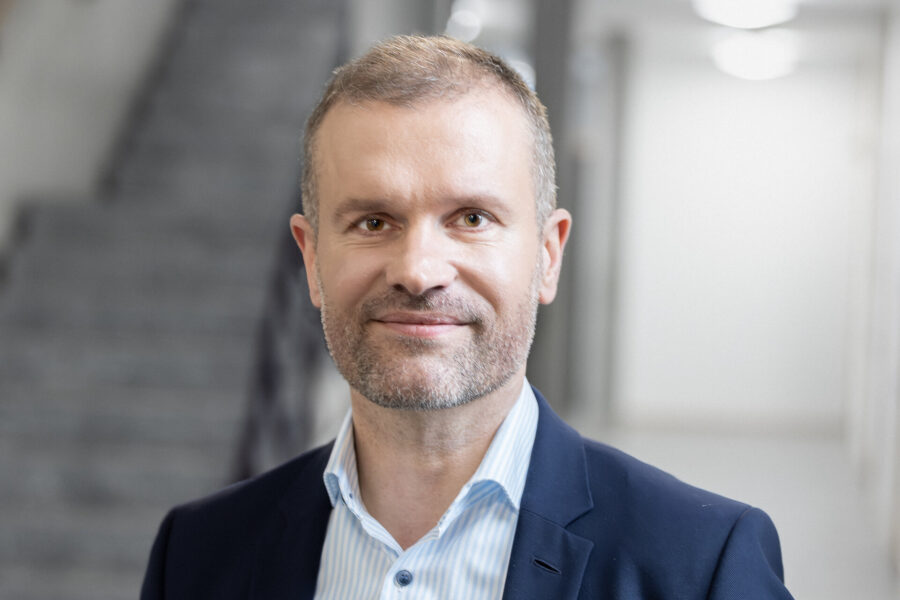 Ein Mann in Anzug und Hemd lächelt in die Kamera, unscharfer Hintergrund.