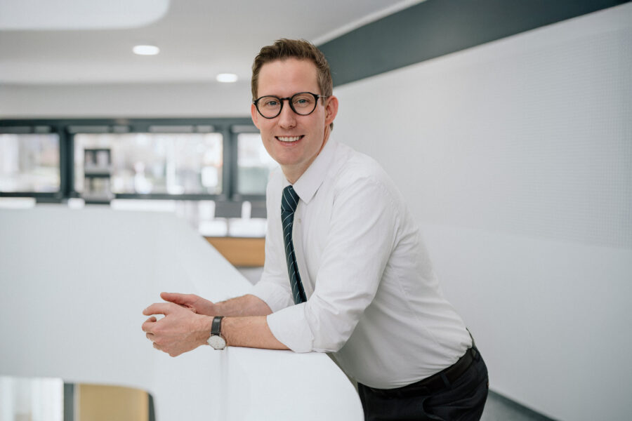 Ein Mann lehnt lächelnd an einem Geländer in einem modernen Innenraum.