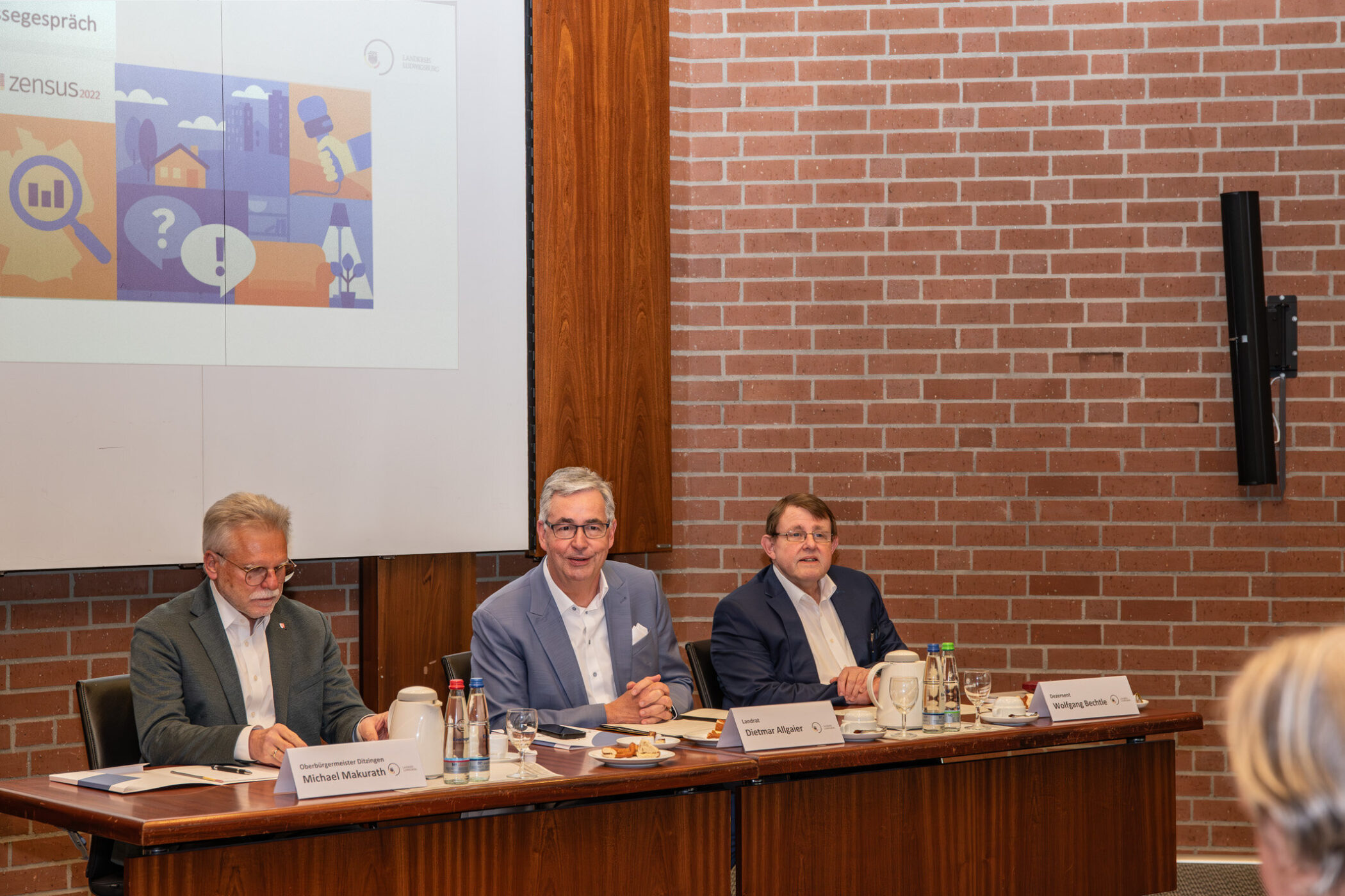 Drei Männer sitzen an einem Tisch in einem Konferenzraum. Präsentation im Hintergrund.