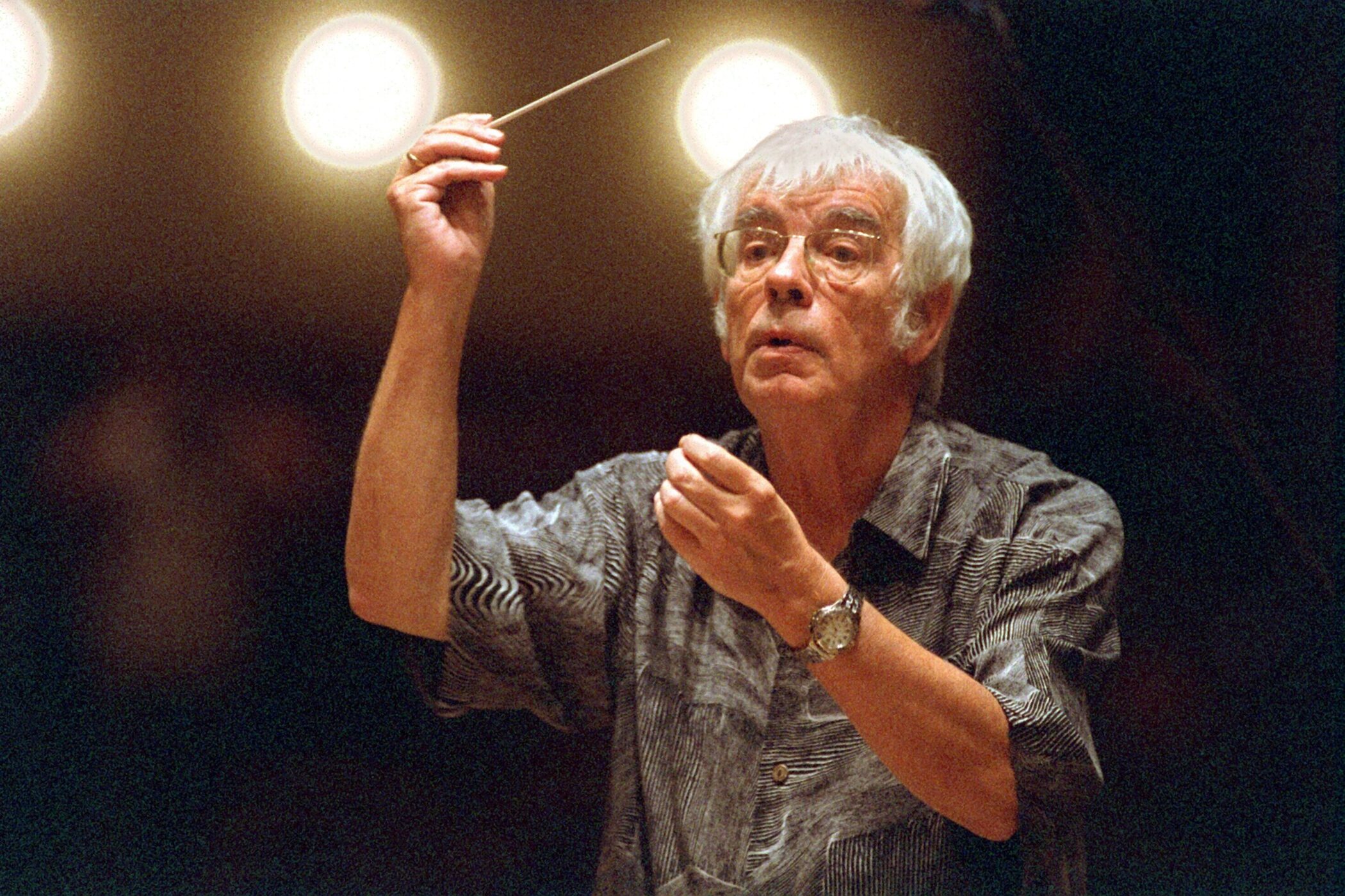 Älterer Mann dirigiert mit Taktstock vor unscharfem Hintergrund.