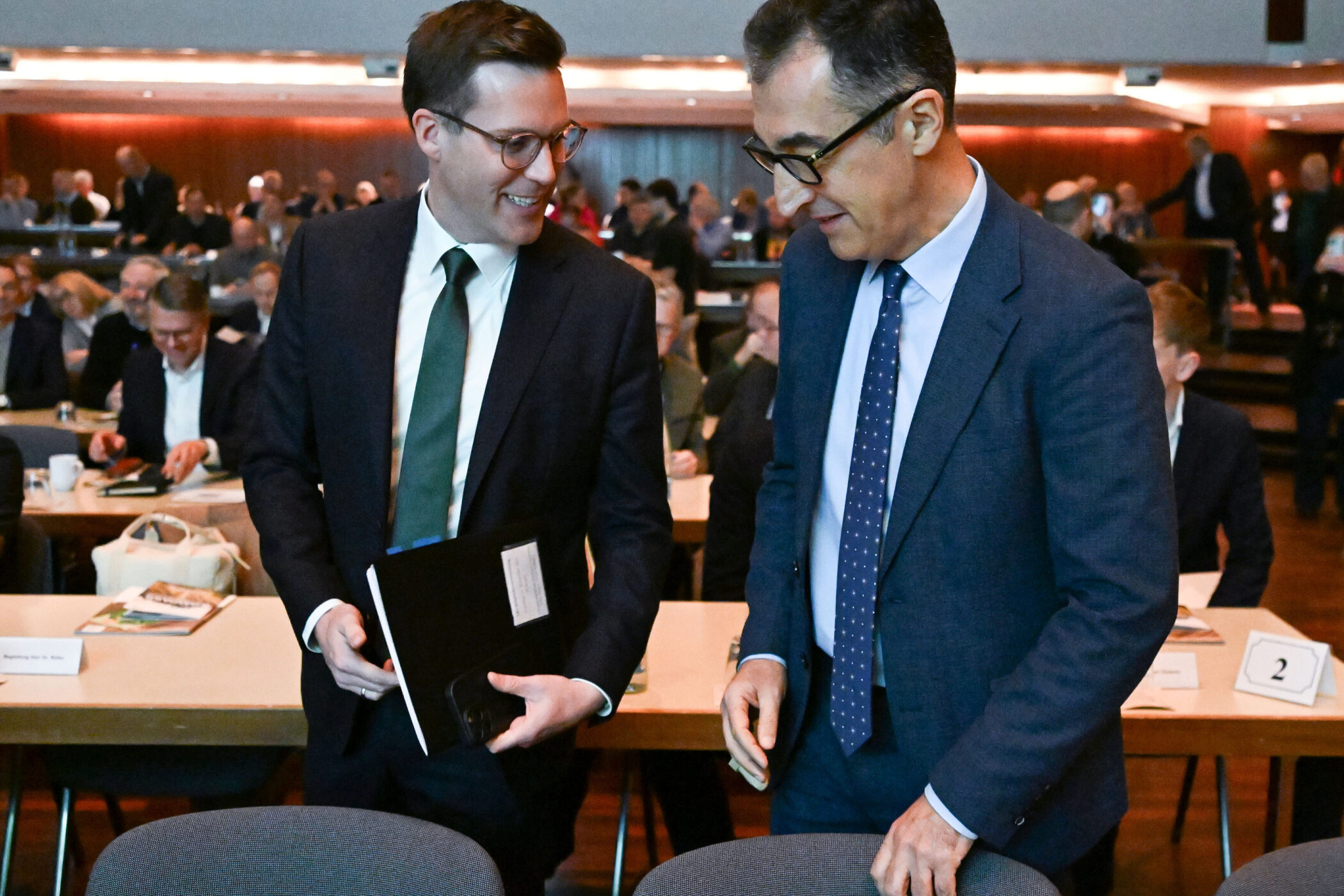 Zwei Männer im Anzug in einem Raum mit Menschen, die an Tischen sitzen.