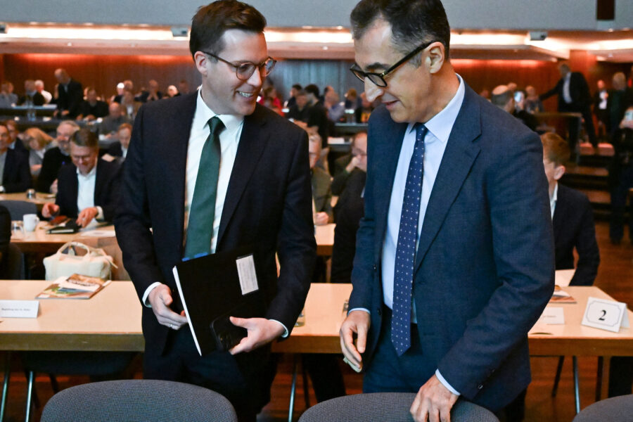 Zwei Männer im Anzug in einem Raum mit Menschen, die an Tischen sitzen.