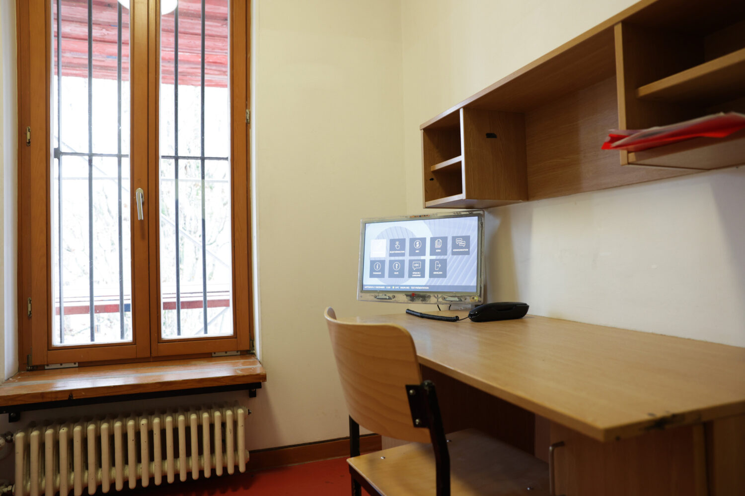 Schreibtisch mit Stuhl, Monitor, Telefon und Regal, Fenster mit Gitterstäben.