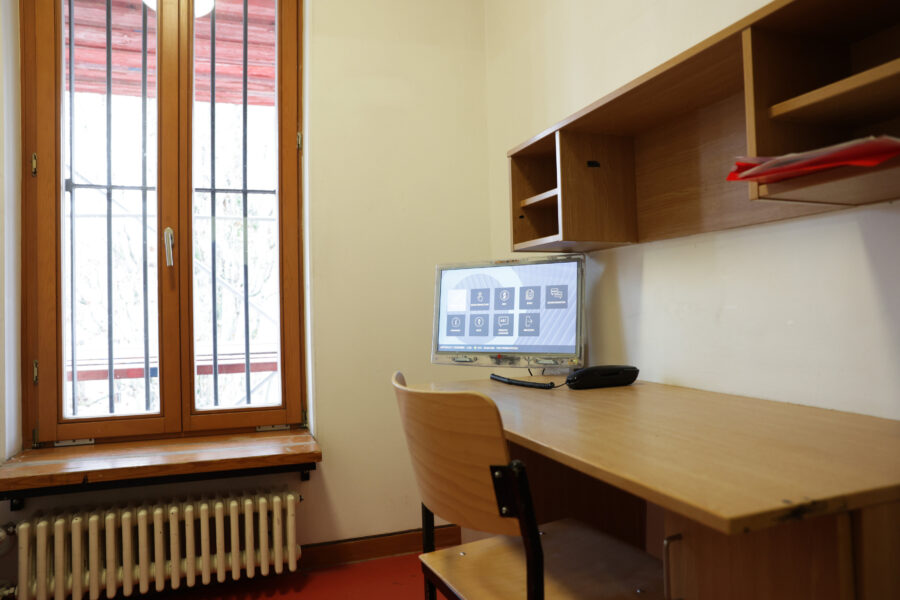 Schreibtisch mit Stuhl, Monitor, Telefon und Regal, Fenster mit Gitterstäben.