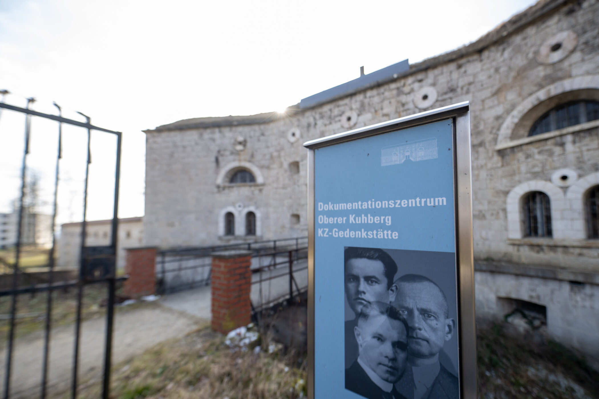 Informationsschild mit Portraits vor Steinbau. Beschriftung: "Dokumentationszentrum Oberer Kuhberg KZ-Gedenkstätte".