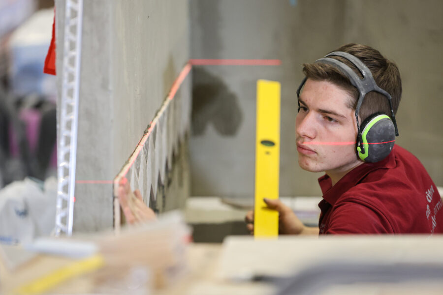 Person mit Gehörschutz hält Wasserwaage an Wand mit Fliesen und Laserlinie.