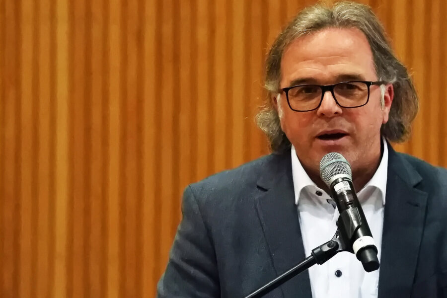 Mann mit Brille spricht in ein Mikrofon vor holzvertäfeltem Hintergrund.