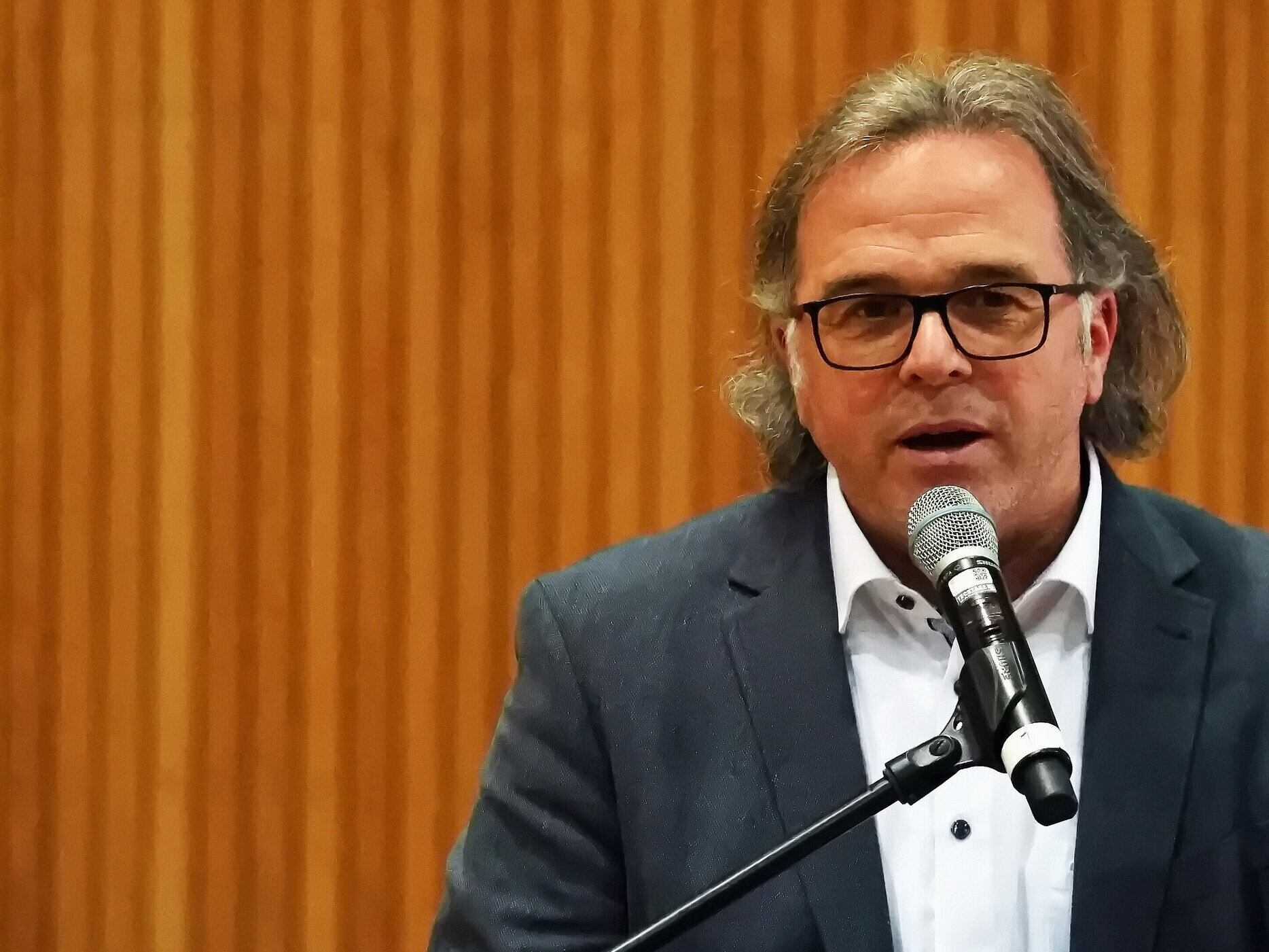 Mann mit Brille spricht in ein Mikrofon vor holzvertäfeltem Hintergrund.