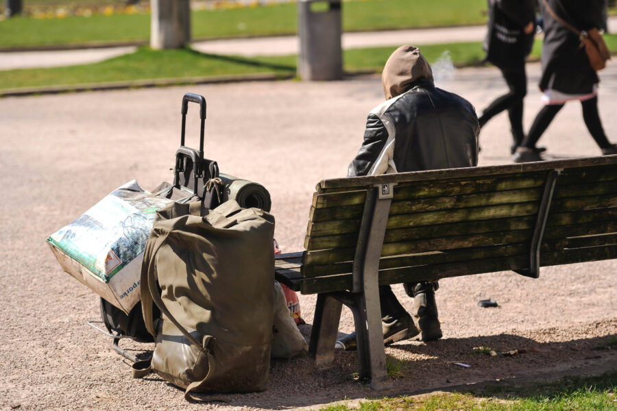 Person sitzt auf Parkbank, Gepäck und Trolley daneben, zwei Personen gehen vorbei.