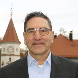 Ein Mann mit Brille vor Gebäuden mit roten Dächern und einem Turm.