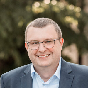 Mann mit Brille und Anzug lächelt, unscharfer Hintergrund mit Bäumen.