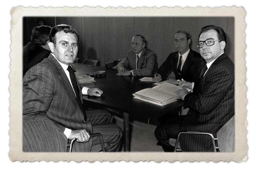 Vier Männer in Anzügen sitzen an einem Tisch mit Papieren. Vintage-Stil.